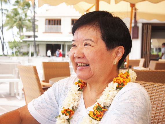 Smiling woman wearing a white floral lei at an outdoor cafe or restaurant.