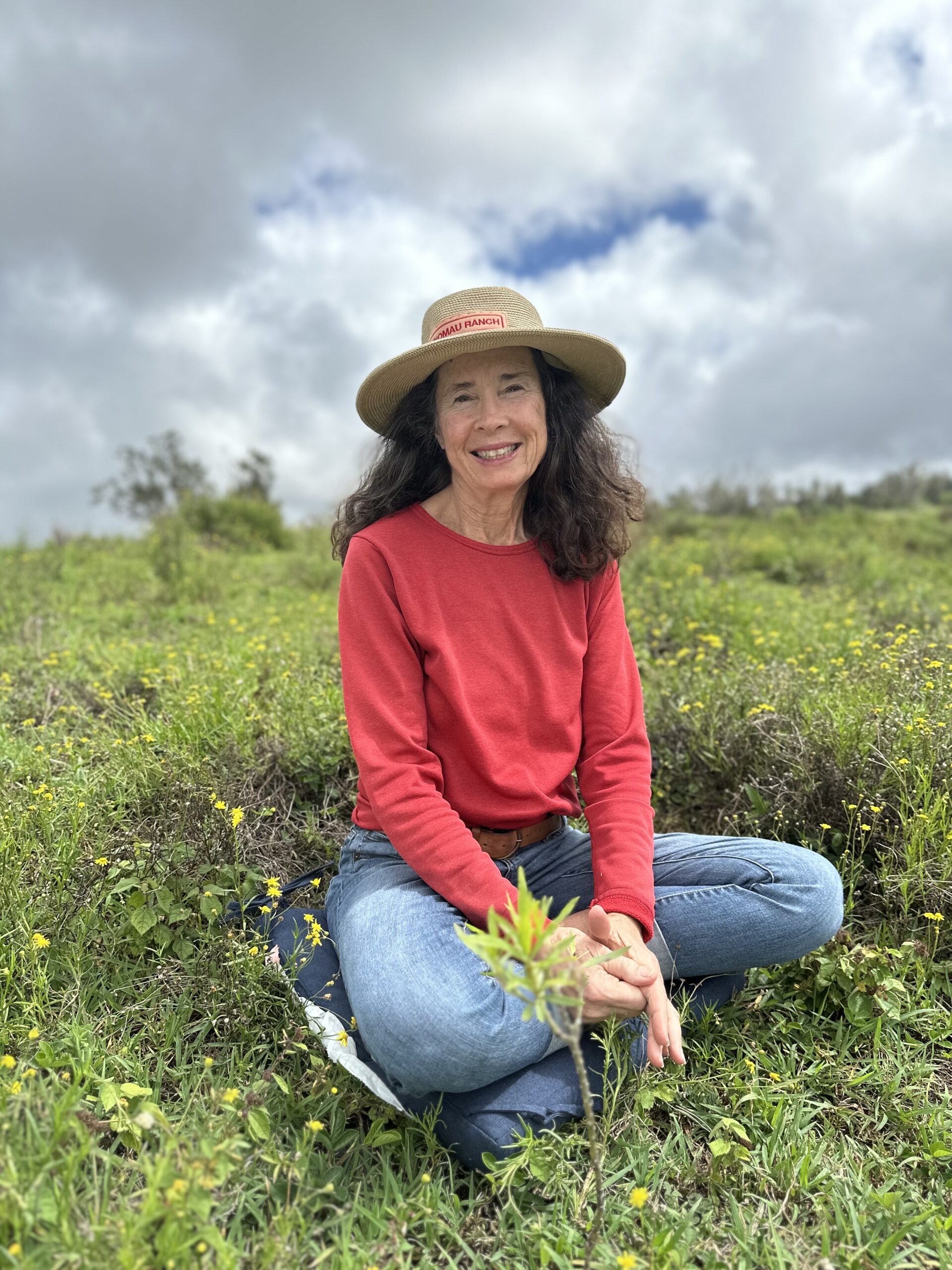 1D1C2613-6730-4B25-B0DF-99F786BAB1A9 Smiling woman sits cross-legged in a meadow of yellow wildflowers, wearing a red shirt and straw hat.