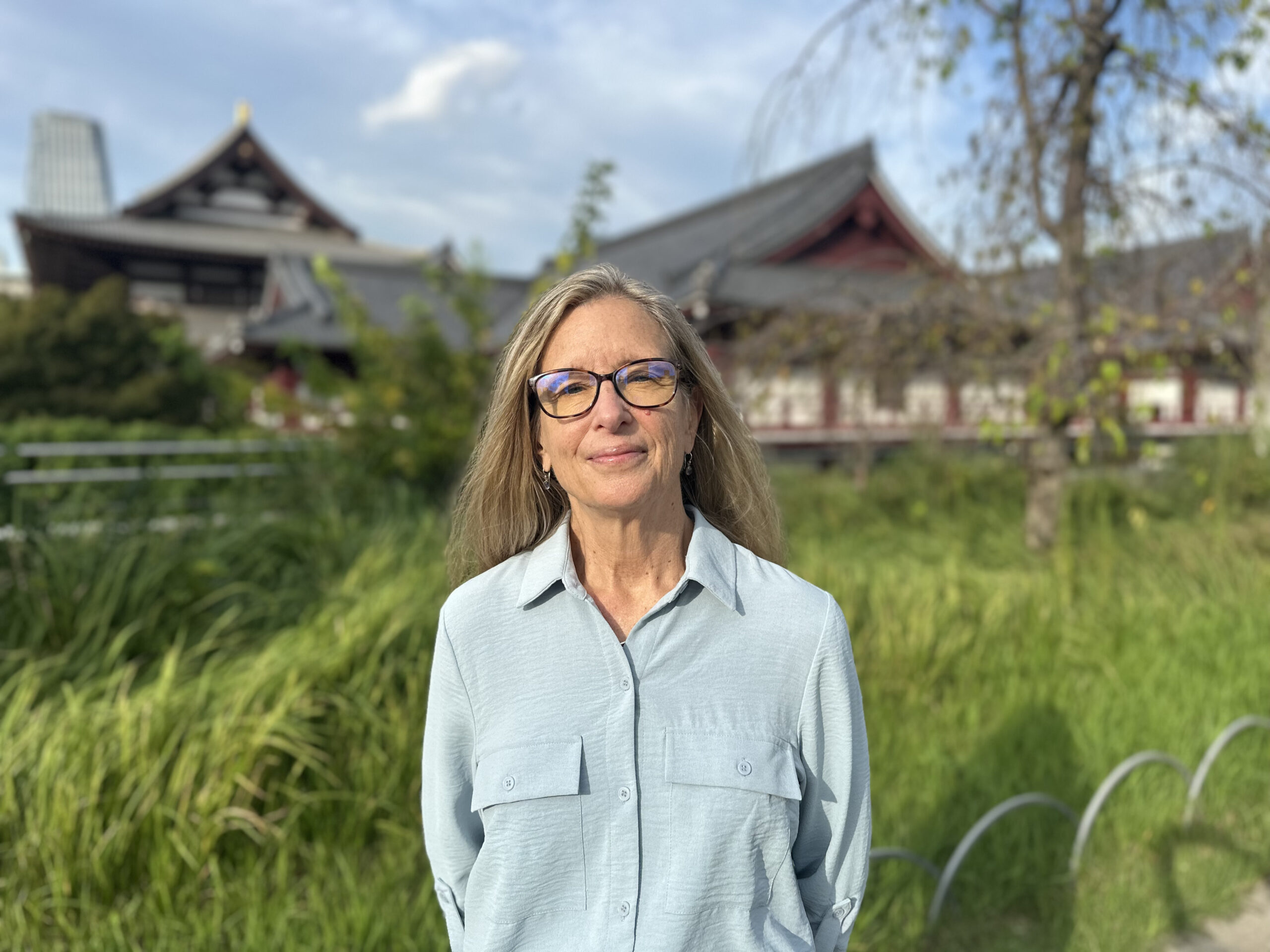 Portrait of a woman with glasses wearing a light blue shirt, standing in a garden with a traditional temple-style building in the background.