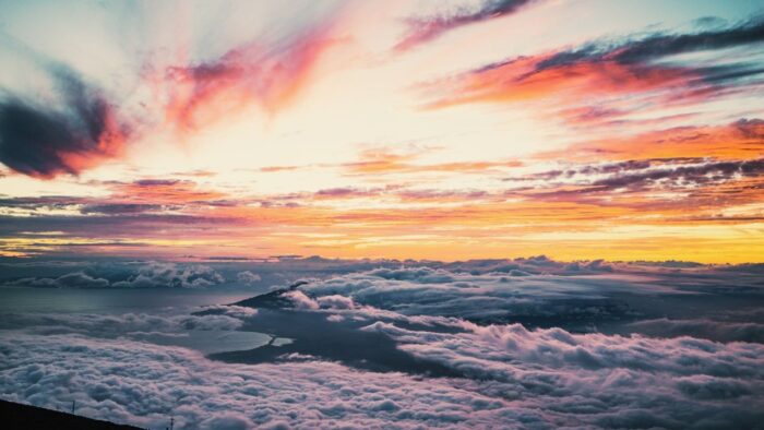 Colorful sunset sky over a blanket of clouds with a distant coastline and island peak visible below.