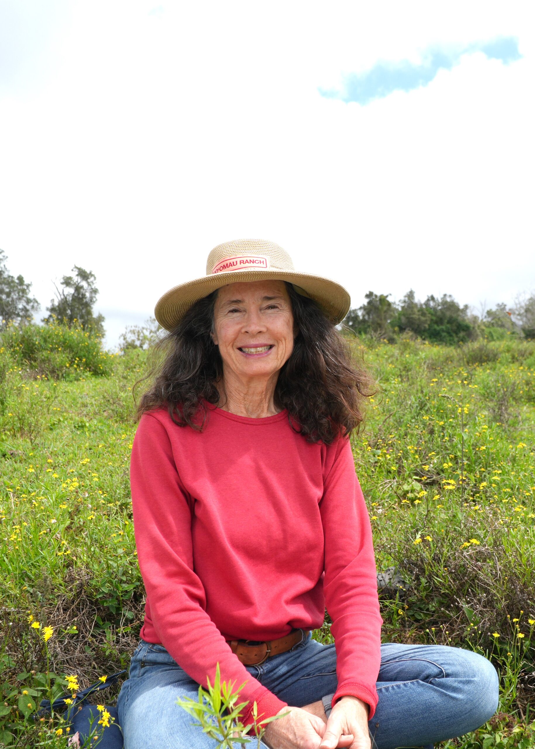 KR_profile_picture Woman wearing a straw hat and red shirt sits in a sunny field of wildflowers, smiling at the camera.